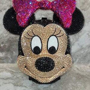 Puede incluir: Un bolso de mano con incrustaciones de pedrer&iacute;a con forma de cabeza de Mickey Mouse. El bolso es negro, dorado y blanco con un lazo rosa en la parte superior.