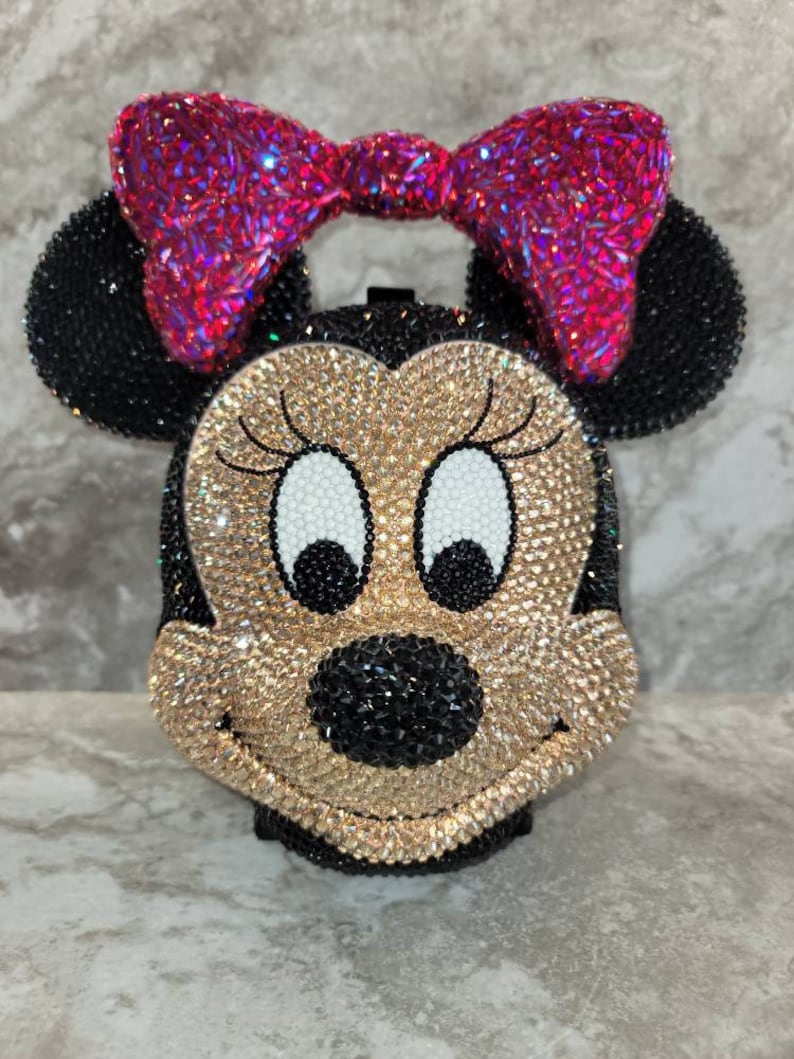 Puede incluir: Un bolso de mano con incrustaciones de pedrer&iacute;a con forma de cabeza de Mickey Mouse. El bolso es negro, dorado y blanco con un lazo rosa en la parte superior.