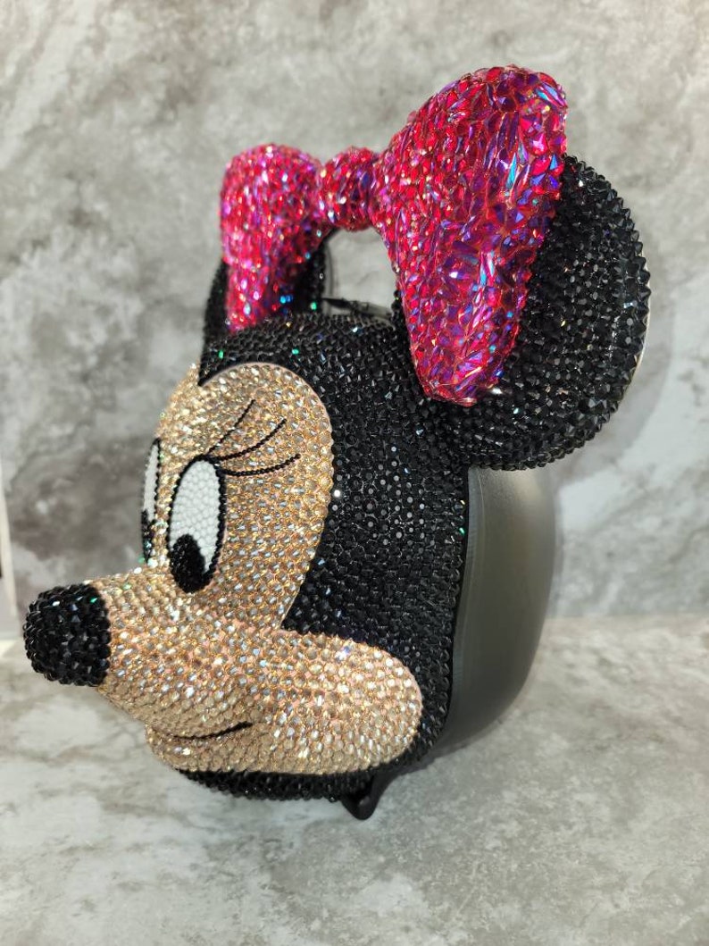 Puede incluir: Un bolso de Minnie Mouse cubierto de pedrer&iacute;a negra y dorada con un lazo rosa.