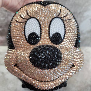 Puede incluir: Un bolso de color negro y dorado cubierto de pedrer&iacute;a en forma de personaje de dibujos animados con ojos grandes y una amplia sonrisa.