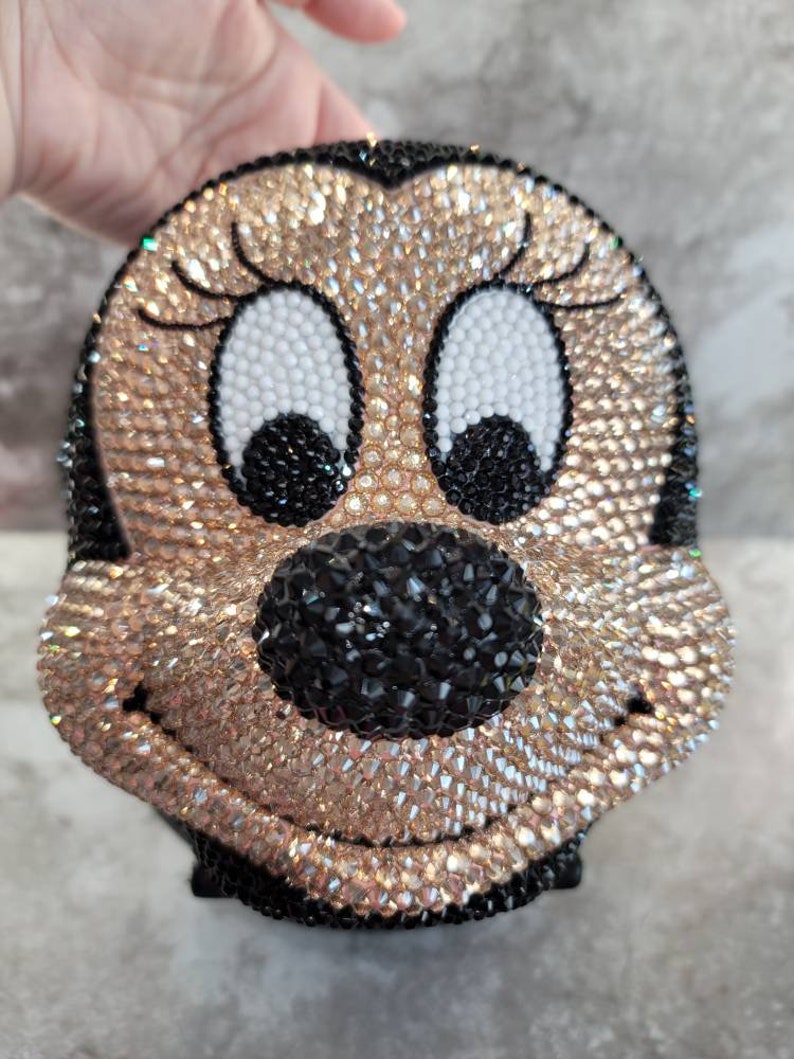 Puede incluir: Un bolso de color negro y dorado cubierto de pedrer&iacute;a en forma de personaje de dibujos animados con ojos grandes y una amplia sonrisa.