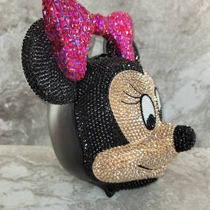 Puede incluir: Un bolso de Minnie Mouse negro y dorado cubierto de pedrer&iacute;a con un lazo rosa.