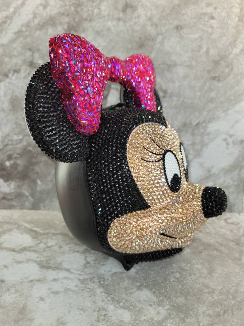 Puede incluir: Un bolso de Minnie Mouse negro y dorado cubierto de pedrer&iacute;a con un lazo rosa.