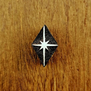 May include: A diamond-shaped pin featuring a black background with a white four-pointed star design centred in the middle. The star has long vertical and horizontal rays that extend towards the edges of the black diamond-shaped surface.