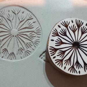 May include: A wooden stamp with a dandelion design and its impression on a grey surface. The stamp is circular, featuring a central point with radiating lines and detailed seed heads. The impression is a mirror image of the stamp.