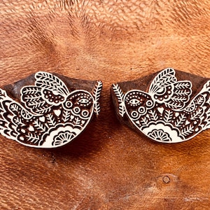 May include: Two wooden stamps with a bird design. The birds are white with intricate patterns and hold a sprig in their beaks. The stamps are set against a brown background.
