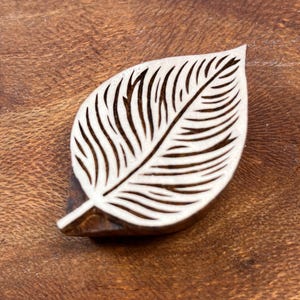 May include: A wooden leaf-shaped stamp with detailed vein patterns. The stamp is light-coloured and rests on a dark brown wooden surface. The leaf design is cut out, creating a negative space effect. Suitable for crafting and decorative projects.