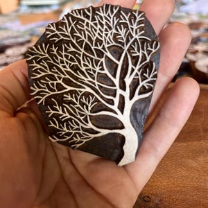 May include: A dark brown, tree-shaped stamp with a white tree design. The stamp is held in a hand, showcasing the intricate details of the tree's branches and trunk. The stamp is likely used for crafting or art projects.