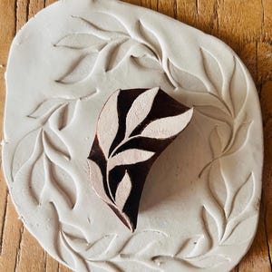 Puede incluir: Un sello de madera marrón oscuro con un diseño de hojas blancas, colocado sobre una impresión de arcilla blanca de hojas. El sello está sobre una base de arcilla blanca con un patrón de hojas, todo sobre un fondo de madera. Suministro de artesanía.