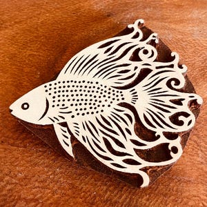 以下が含まれることがあります： 詳細な、様式化された魚のデザインが施された木製のブロック。 魚は大きく流れる尾とひれを持ち、体は小さな点で覆われています。 デザインは、濃い茶色の背景に対して明るい色で彫られています。