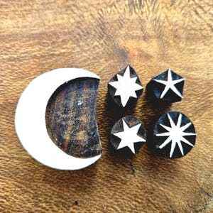 May include: Wooden stamps featuring celestial designs. A crescent moon stamp and four star stamps in various shapes are displayed. The stamps have white designs on a dark wood base, perfect for crafting and adding a celestial touch to projects.