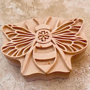 以下が含まれることがあります： 詳細な彫刻が施された木製の蜂のスタンプ。このスタンプは、複雑な羽と体の模様を持つ蜂が特徴です。木材は自然な淡い茶色で、工芸品や装飾目的のために設計されています。