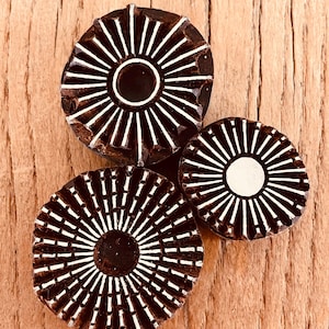 May include: Three dark brown, circular wooden stamps with white radial designs. The stamps vary in size, with a central circle and radiating lines. The stamps are arranged on a light brown wooden surface.