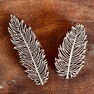 May include: Two wooden feather-shaped stamps with intricate white line details. The stamps are made of dark brown wood and are set against a textured, brown wooden surface. These stamps are likely used for textile or paper printing.