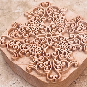 Könnte beinhalten: Ein Holzstempel mit einem detaillierten, symmetrischen floralen Design, das in seine Oberfläche geschnitzt ist. Der Stempel ist quadratisch mit abgerundeten Ecken und weist komplizierte Wirbelmuster und florale Motive auf. Das Holz hat eine natürliche, hellbraune Farbe.