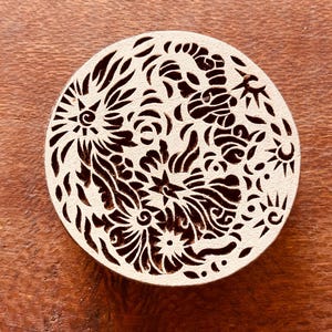 以下が含まれることがあります： 花とスターバーストの複雑な切り抜きデザインが施された、丸いクリーム色のコースター。コースターは明るい色の素材でできており、木製の表面に置かれています。切り抜かれたパターンは、装飾的で風通しの良い美しさを生み出しています。