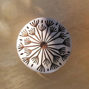Könnte beinhalten: Ein runder Holzblock mit einem geschnitzten Design eines Löwenzahns. Das Design ist in Schwarzweiß und zeigt einen Kreis in der Mitte mit strahlenden Linien.