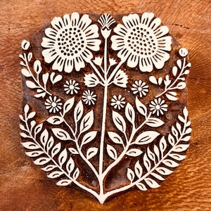 Puede incluir: Un sello de madera marrón para estampado con un diseño floral simétrico. El tallado muestra dos grandes flores similares a girasoles en la parte superior, flanqueadas por flores más pequeñas y tallos frondosos que descienden hacia una base puntiaguda.