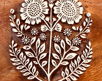May include: A brown wooden block print stamp featuring a symmetrical floral design. The carving displays two large sunflower-like blooms at the top, flanked by smaller flowers and intricate leafy stems extending downwards to a pointed base.