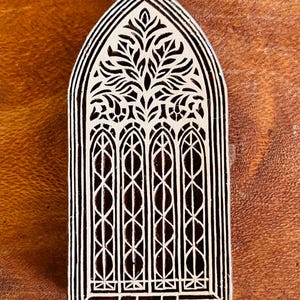 May include: A wooden block with a carved design resembling a cathedral window. The design features a central tree-like motif above four arched window panels with intricate lattice patterns. The block is light-colored against a dark wood surface.
