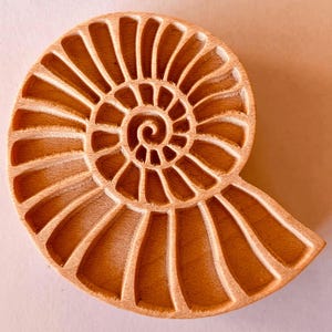 Könnte beinhalten: Ein dekoratives Objekt aus Holz in Form eines Nautilus. Das hellbraune Holz ist in einem Spiralmuster mit detaillierten Rippen geschnitzt. Das Design ist eine natürliche, organische Form. Die Holzmaserung ist sichtbar und trägt zur Ästhetik des Objekts bei.