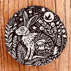 Puede incluir: Posavasos redondo en blanco y negro con un conejo rodeado de follaje, estrellas y una luna con silueta de pájaro. El diseño es una ilustración de estilo linograbado, para decoración.