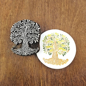 May include: A dark brown wooden printing block featuring an intricate tree of life design with birds nestled in the branches, positioned next to a white circular coaster displaying a stamped version of the same tree in yellow, green, and brown ink.
