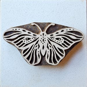 May include: A wooden butterfly block with a detailed white design. The butterfly has intricate wing patterns and antennae. The block is rectangular with a dark brown, aged appearance, and is likely used for printing or stamping.