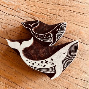 May include: Two wooden whale-shaped stamps. The larger whale is white with black lines and star details. The smaller whale is black with white accents. The stamps are on a wooden surface.