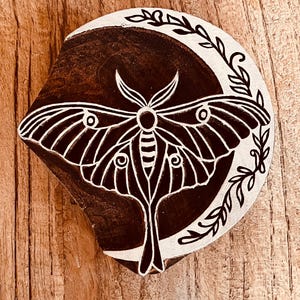 May include: A wooden block featuring a moth and crescent moon design. The moth is white with detailed wing patterns, set against a dark brown background. A white crescent moon shape surrounds the moth, with leafy accents. The block is on a wooden surface.