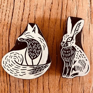 May include: Two carved wooden printing blocks featuring black and white relief designs. One block depicts a curled fox with detailed fur texture, while the other block shows an upright hare with long ears and patterned markings on its body.