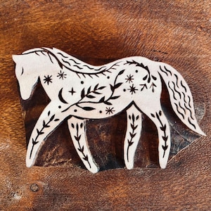 Puede incluir: Un sello de madera para estampar con la silueta de un caballo. El caballo está decorado con patrones grabados en negro de hojas, enredaderas, estrellas y una luna creciente. El sello tiene una superficie clara en relieve sobre madera oscura.