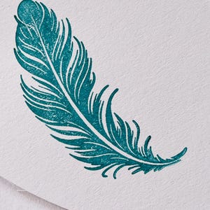 May include: A teal feather stamp on white paper. The stamp shows detailed lines and curves, creating a realistic feather design. The image is a close-up, highlighting the texture of the paper and the stamp's intricate details.