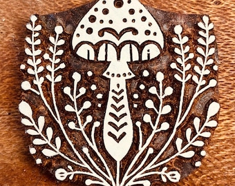 May include: A wooden stamp with a mushroom design, featuring a dotted cap and a chevron-patterned stem. The mushroom is surrounded by white floral elements. The stamp is set against a brown wooden surface.