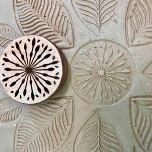 May include: A close-up of a wooden block with a carved design of a sunburst. The block is resting on a light brown surface with a pattern of leaves and sunbursts.