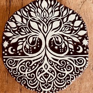 Puede incluir: Un bloque de impresión circular de madera con un diseño profundamente tallado de un árbol con ramas y raíces intrincadas. La superficie elevada está cubierta de pintura blanca, contrastando con la madera marrón oscura de las áreas.