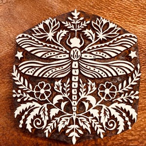 May include: A wooden block with a white dragonfly design. The dragonfly is centered, with detailed wings and body. Floral and leaf patterns surround the dragonfly, with two small stars. The block is a dark brown color.