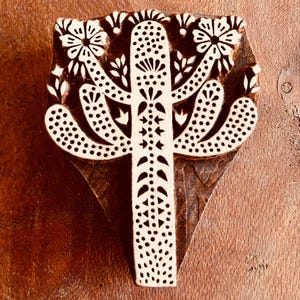Puede incluir: Un sello de impresión de bloque de madera con un diseño detallado de cactus. El sello tiene una forma de cactus beige claro con intrincados patrones florales y de puntos, sobre un fondo marrón oscuro. El diseño es simétrico y adecuado para la impresión textil o en papel.