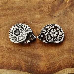May include: Two wooden printing blocks shaped like hedgehogs, each featuring a detailed black and white floral pattern carved into the surface. The designs depict a hedgehog with a prominent flower on its back, surrounded by intricate leaf motifs.