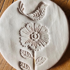 May include: A round, off-white ceramic piece featuring an embossed design. The design includes a stylized bird at the top and a detailed flower with leaves below. The piece is likely a decorative element or a craft supply.