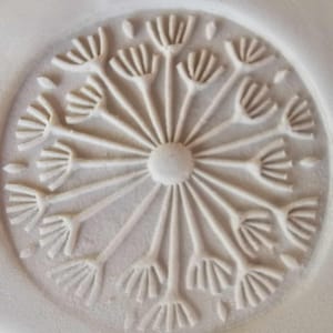 Peut inclure: Une sculpture en relief circulaire et blanc cassé d'un capitule de pissenlit. Le motif présente une sphère centrale avec des tiges rayonnantes se terminant par des grappes de graines détaillées. La sculpture est sur un fond texturé blanc cassé.