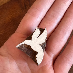 May include: A small, rectangular pendant featuring a white hummingbird design on a dark brown background. The hummingbird is in flight, with its wings spread. The pendant is held in a person's hand.
