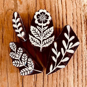May include: Four wooden block stamps with botanical designs. The stamps feature white floral and leaf patterns against a dark brown background. The blocks are arranged on a wooden surface.