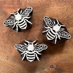 May include: Three wooden bee stamps with white bee designs. The bees have detailed wings and striped bodies. The stamps are on a wooden surface, showcasing the natural wood grain.