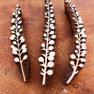 May include: Three wooden printing blocks with a botanical design. Each block is a dark brown color with a white floral pattern. The blocks are arranged on a wooden surface.