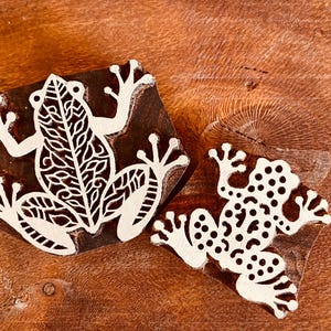 May include: Two wooden frog stamps with white frog designs. One stamp features a detailed frog with leaf-like patterns, while the other has a dotted frog design. The stamps are set against a warm, brown wooden surface.