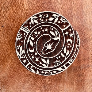 May include: A circular, dark brown wooden coaster with a white celestial design. The design includes a spiral, stars, moons, suns, and leaves. The coaster is set against a brown wooden surface.