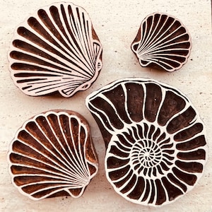 May include: Four wooden seashell stamps with white and brown details. The stamps feature various shell designs, including a spiral nautilus and several scallop shells. These stamps are ideal for crafting and art projects.