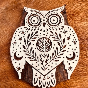 May include: A wooden block print stamp featuring a detailed owl design. The owl is carved with intricate patterns including stars, crescent moons, and leafy branches. The light-coloured wood surface contrasts with the dark, recessed carved lines.