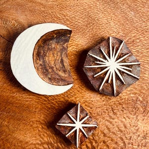 May include: Three wooden stamps with celestial designs: a crescent moon, a starburst, and a diamond-shaped star. The stamps are brown with white accents and are arranged on a wooden surface.